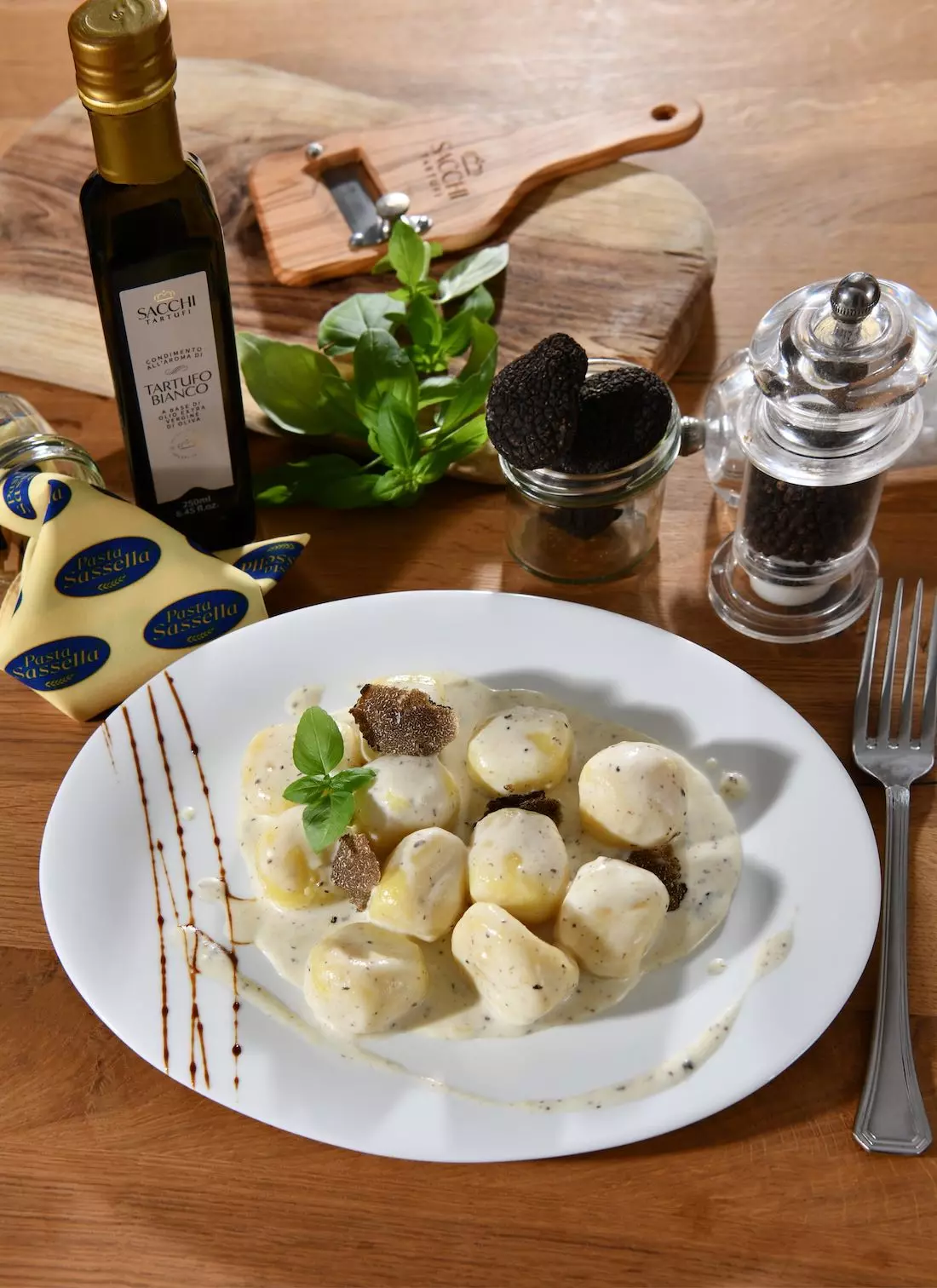 Gnocchi in cremiger Sauce, garniert mit frischen schwarzen Trüffelscheiben und Basilikum. Im Hintergrund stehen Trüffelöl, ganze Trüffel und ein Pfefferstreuer.
